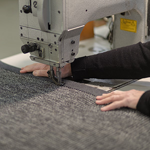 Maßarbeit beim Einfassen. Foto von einer Nähmaschine mit der gerade ein Teppich mit einer Bordüre eingefasst wird.